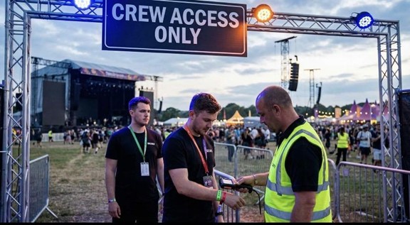 A security worker in a yellow vest checks the credentials of a man under a Crew Access Only sign at an outdoor event, with a crowd and stage visible in the background.