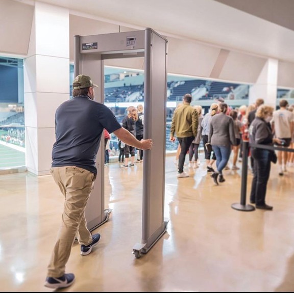 A man pushes a walk-through metal detector on wheels while people line up to enter a stadium in the background. The scene is indoors, with a view of a sports field through open windows.
