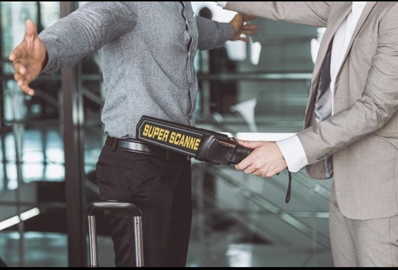 A security officer in a suit uses a handheld metal detector on a person with arms outstretched during a security check; the person has a suitcase nearby.