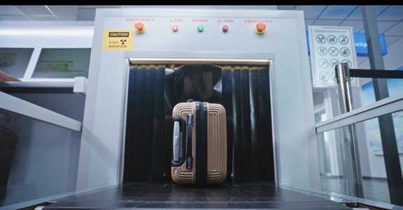A brown suitcase passes through an airport security X-ray scanner, which displays buttons and warning signs above the conveyor belt.