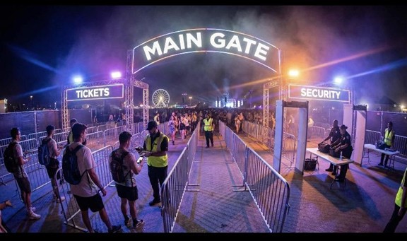 A brightly lit MAIN GATE arch welcomes crowds at an outdoor event at night. People line up for tickets and security checks, with staff guiding them and a ferris wheel visible in the background.