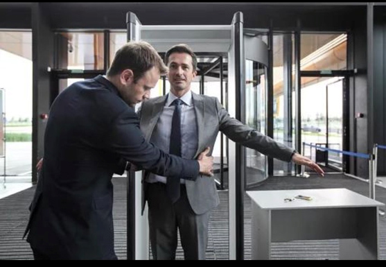 A security officer in a suit conducts a pat-down search on another man in a suit, who stands with arms outstretched at a security checkpoint with a metal detector gate and a tray containing keys on a table nearby.