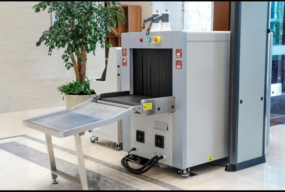 An X-ray baggage scanner machine in an indoor setting, with a conveyor belt and metallic tray for luggage, positioned next to a potted plant.