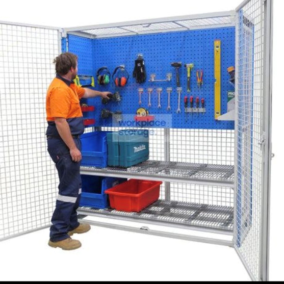 A man in a high-visibility shirt stands in front of a metal storage cage with shelves and a blue pegboard, organizing tools and safety equipment. Red and blue plastic bins are on the shelves below.