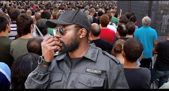 A security guard in uniform and sunglasses speaks into a walkie-talkie while facing a large crowd of people gathered near a metal fence.