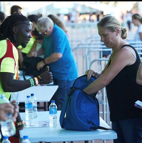A security staff member gives a thumbs up as a woman opens her blue bag for inspection at a checkpoint table, with other people and bottles of water visible in the background.