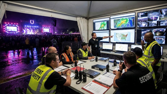 Event safety and security personnel sit at a table under a tent, monitoring weather maps and security camera feeds on large screens, while a concert takes place on a brightly lit stage in the background at night.