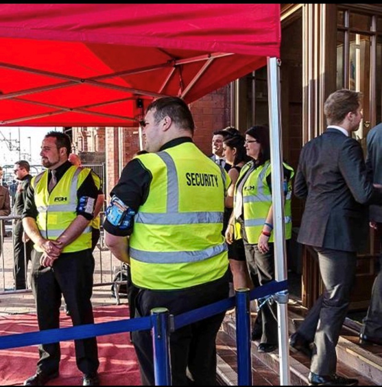 Security personnel in yellow vests stand under a red canopy by a building entrance, monitoring an event as people in formal attire line up outside. A blue rope barrier separates the entrance area.