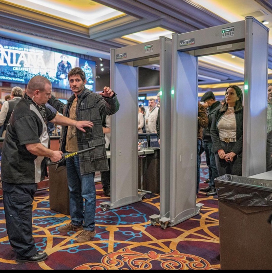 A security guard uses a handheld scanner on a man passing through a metal detector; a woman waits her turn nearby. People and event signage are visible in the background.
