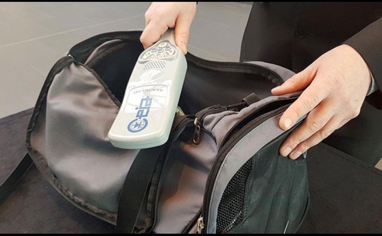 A person holds a handheld metal detector over an open black and gray bag, inspecting its contents on a flat surface.