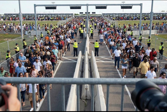 Large crowds of people walk through fenced lanes monitored by security personnel wearing yellow vests at an outdoor event or venue, with clear skies above and cameras visible in the foreground.